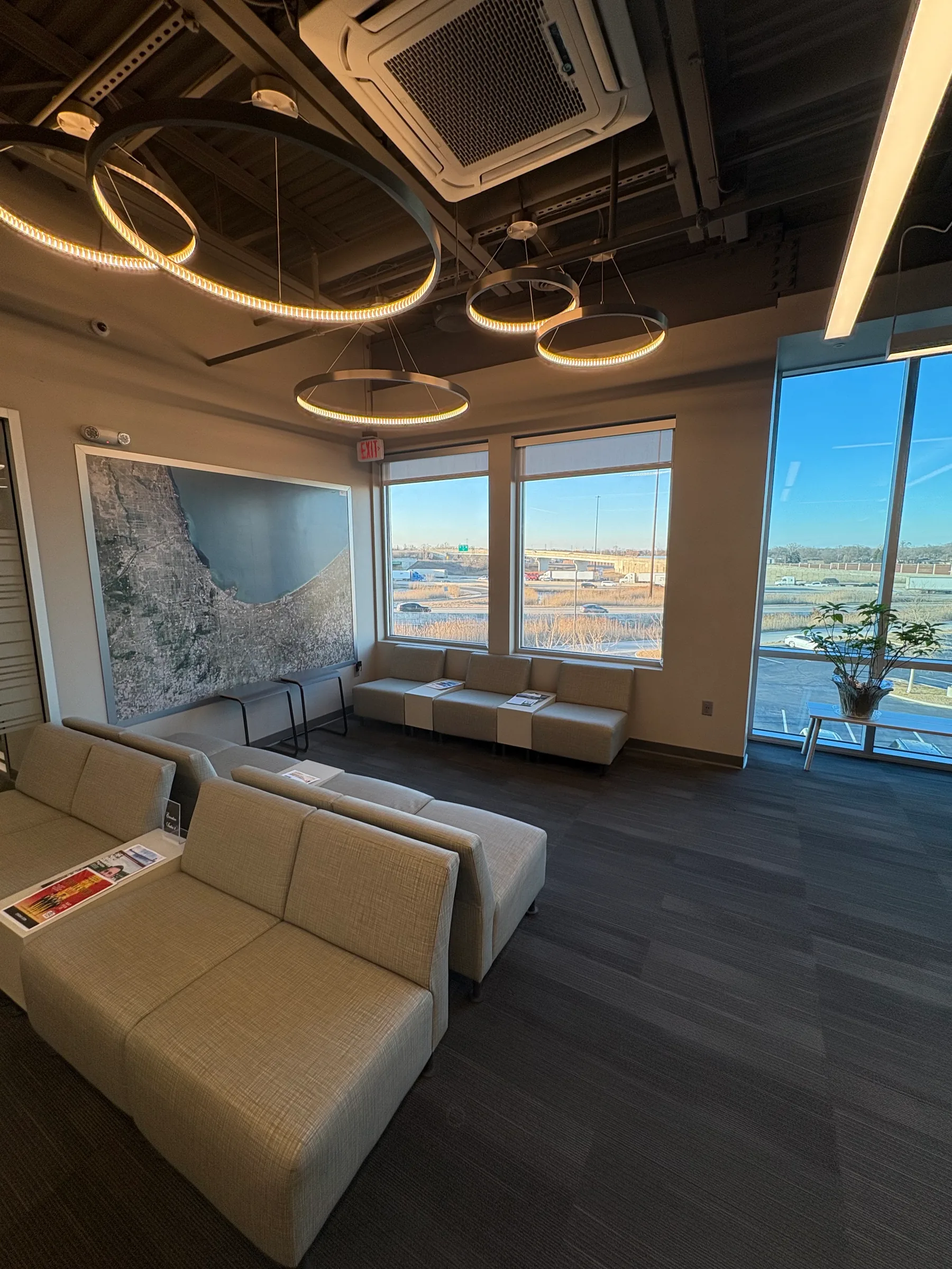 Modern office lounge with beige seating, pendant lighting, and waterfront views through large windows.