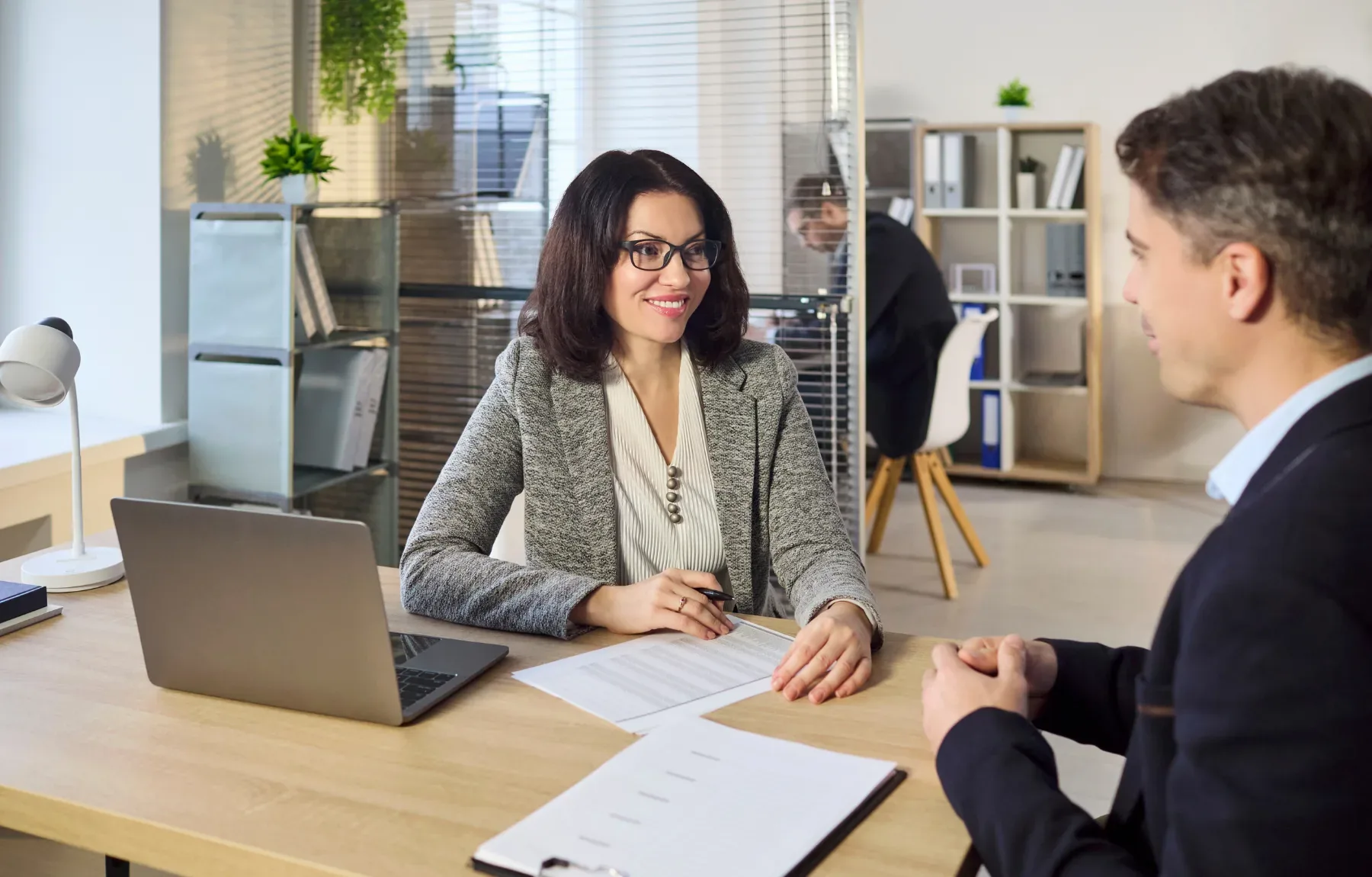 A smiling woman with dark curly hair and glasses, wearing a gray blazer, sits at a wooden desk across from a man in a dark su