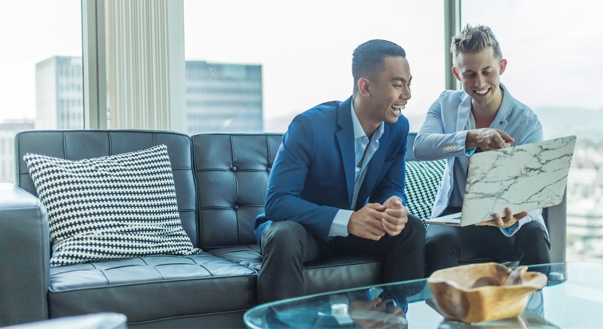 Two business professionals review architectural plans on a leather sofa in a high-rise office.